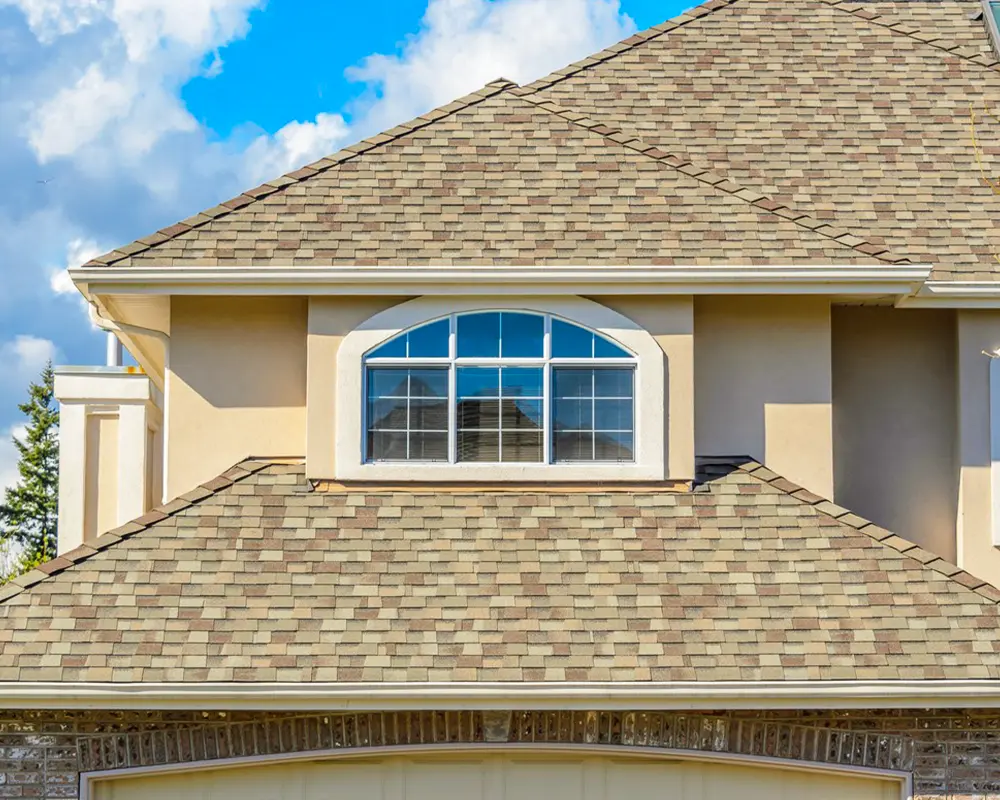 Close-up of a tan house with a freshly installed shingle roof under a bright blue sky, highlighting durability and style.