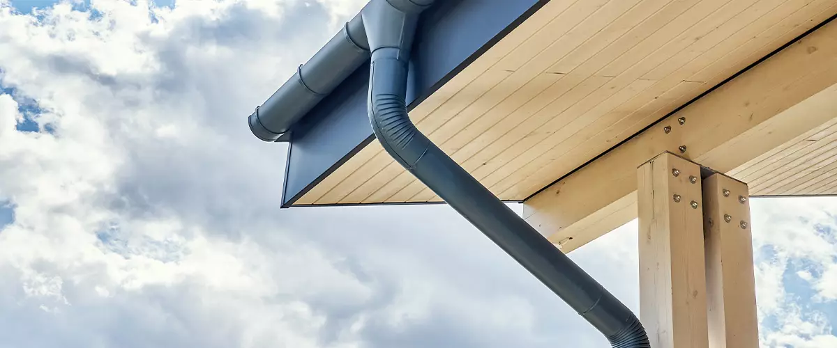 Contemporary grey metal rainwater downpipe installed on roof of new building with wooden terrace on cloudy day close view